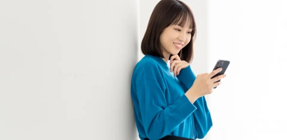 壁に背を預けて笑顔でスマートフォンを操作している女性